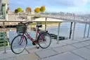 Red bicycle with a basket leaning on a railing by a waterfront path and marina