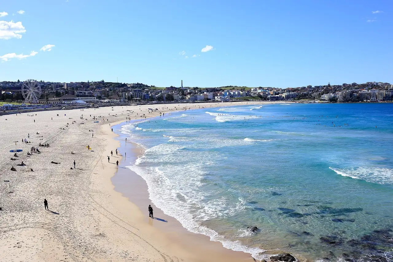 Long curve of Bondi Beach with pale sand, turquoise surf and houses along the shore on a sunny day