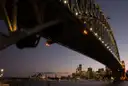 The steel arch of the Sydney Harbour Bridge seen from below at dusk, with the city lights beyond