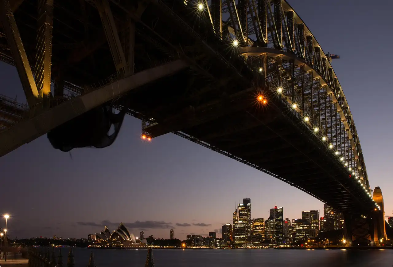 The steel arch of the Sydney Harbour Bridge seen from below at dusk, with the city lights beyond