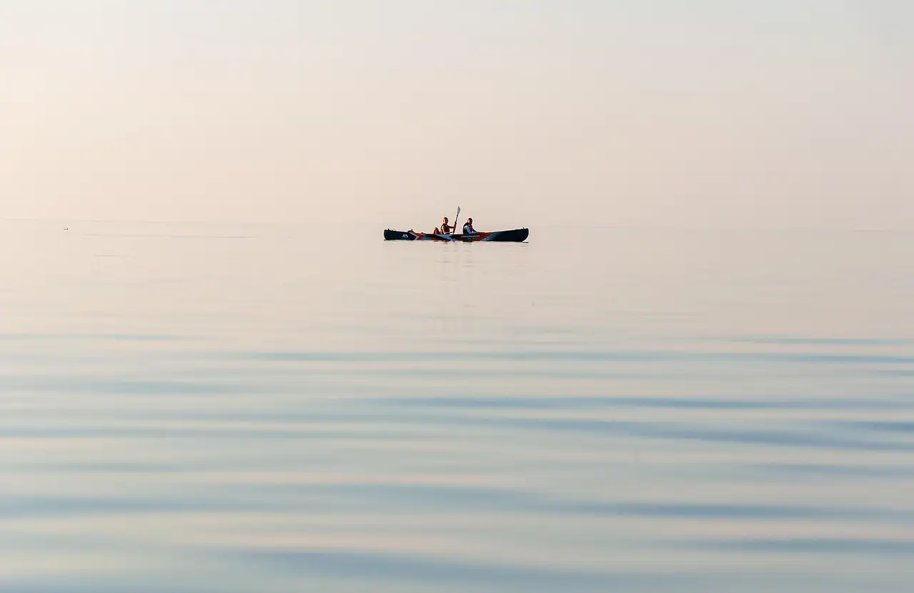 A kayak with two paddlers far out on still water in soft morning light