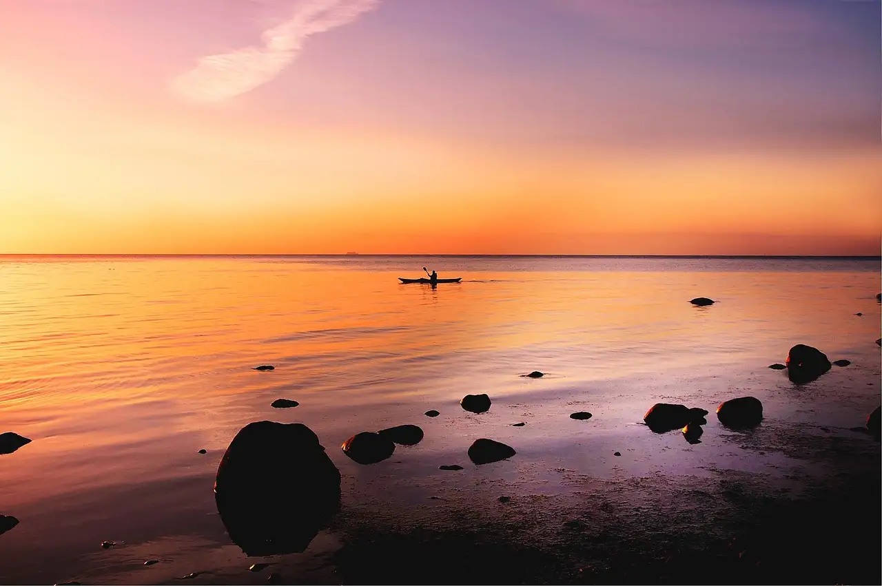 A small kayak on calm water near a rocky shore, with the sun low on the horizon