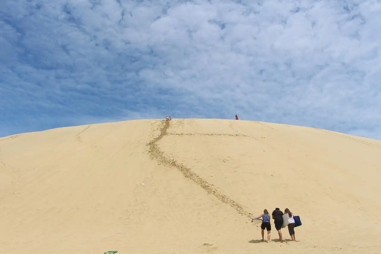 A pale sand dune under a cloudy sky, with three walkers far off at its foot