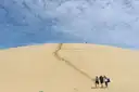 A pale sand dune under a cloudy sky, with three walkers far off at its foot