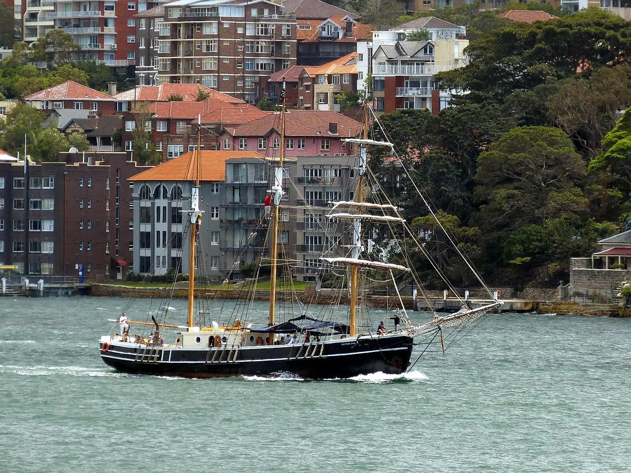 A two-masted sailing ship on Sydney Harbour in front of waterfront houses