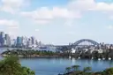 Sydney Harbour seen from a green headland, with sailing boats, the city skyline and the Harbour Bridge in the distance