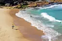 A surfer carrying a board along a sandy beach next to turquoise waves