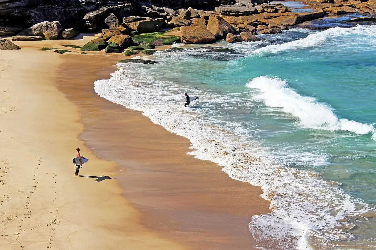 A surfer carrying a board along a sandy beach next to turquoise waves