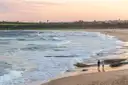 Two people walking along a Sydney beach at sunset beside rolling waves
