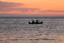 Two people paddling a kayak on calm sea water at sunset, seen as dark shapes against a pink sky