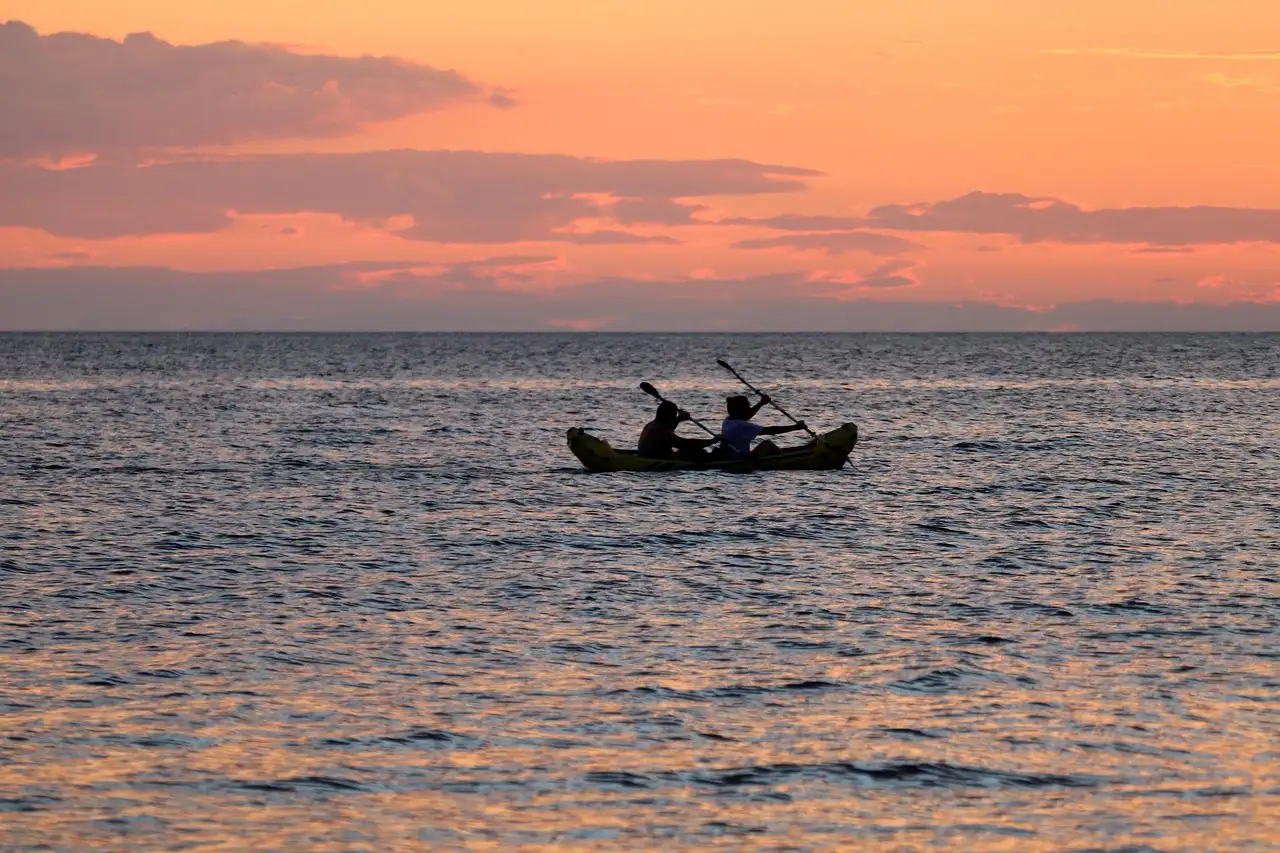 Two people paddling a kayak on calm sea water at sunset, seen as dark shapes against a pink sky
